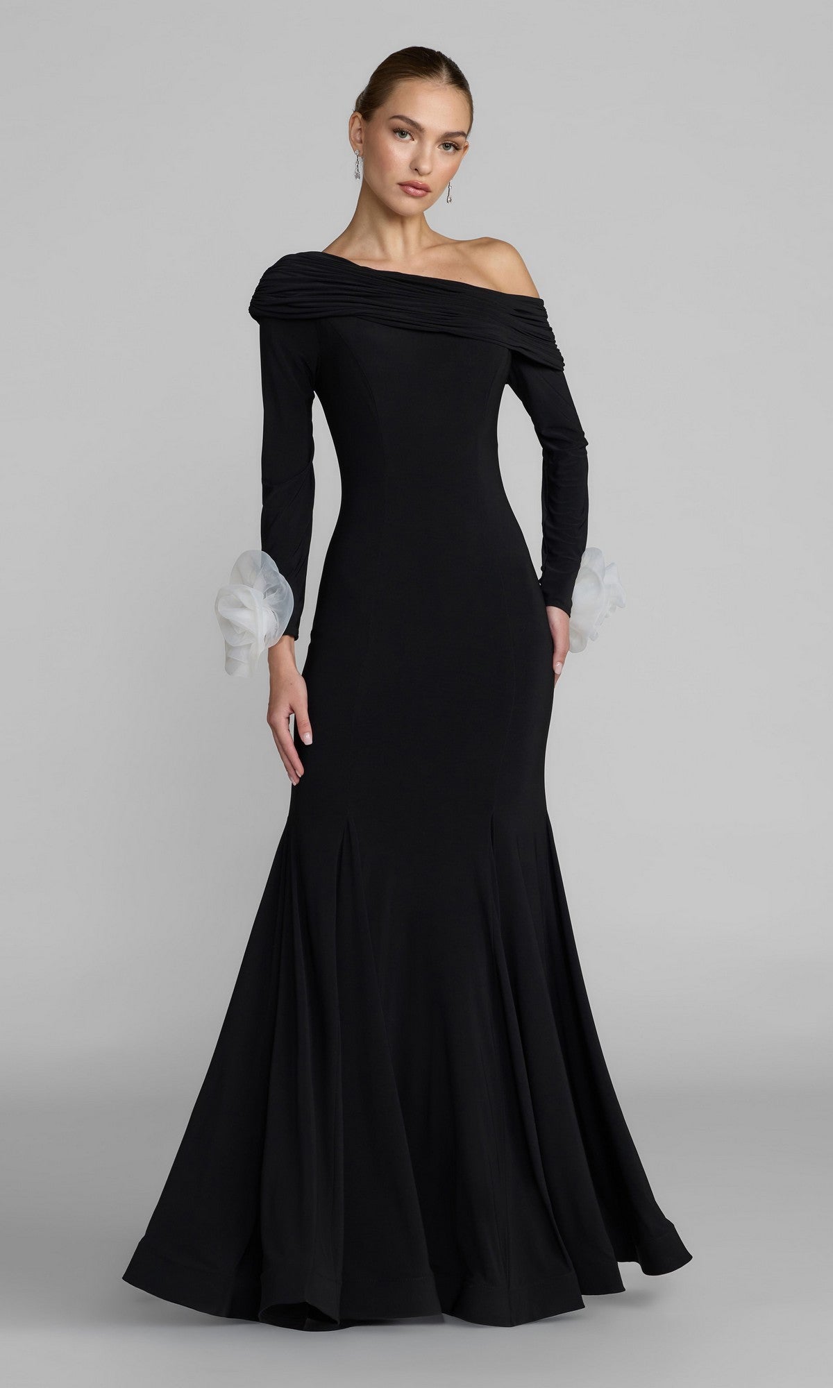 A woman wearing the Long Formal Dress: Mac Duggal 50134 in Black Ivory, featuring a long, fitted mermaid silhouette with an off-the-shoulder neckline, flared hem, and sheer white ruffled cuffs, stands against a plain gray background.